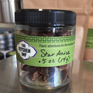 May include: A glass jar filled with brown star anise, labeled "The Kitchen Imp" and "Star Anise 5 oz (149)". The label is green with white text and a purple border.