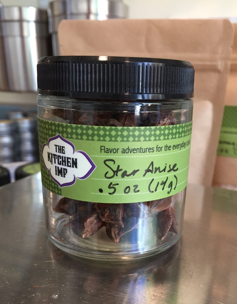 May include: A glass jar filled with brown star anise, labeled "The Kitchen Imp" and "Star Anise 5 oz (149)". The label is green with white text and a purple border.