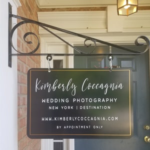 May include: Black sign with gold trim hanging from a black metal bracket. The sign reads "Kimberly Coccagna Wedding Photography New York | Destination www.kimberlycoccagna.com By Appointment Only".