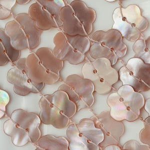May include: A collection of light pink, flower-shaped buttons made from iridescent shell material. Each button features two holes in the center and is strung together in rows using a thin, light-colored thread, creating a repeating pattern.