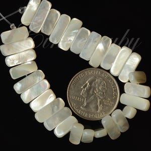 May include: A strand of rectangular, iridescent mother-of-pearl beads. A U.S. quarter coin is next to the beads for size comparison. The beads are strung on a thin, clear string against a black background. The beads have a pearly, opalescent sheen.