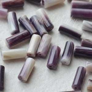 以下が含まれることがあります： 紫と白のストライプ模様の貝殻ビーズの集まり。ビーズは円筒形で、滑らかで磨かれた仕上げになっています。