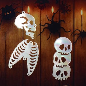 May include: White iridescent skull and rib cage pendants for Halloween. The pendants are made of a pearlescent material and are designed to be strung. The background features spiders and candles.