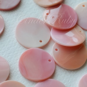 May include: A collection of pink, round, iridescent shell sequins with a small hole in the center of each. The sequins are arranged on a white surface.
