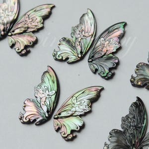 May include: Close-up of several iridescent butterfly wing charms. The wings feature a detailed, carved design with a mix of colors, including green, pink, and silver. These decorative charms are ideal for jewelry making or craft projects.