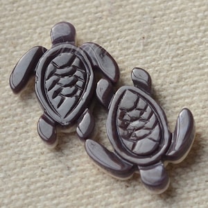 May include: Two dark purple sea turtle shaped glass buttons. The buttons are detailed with a textured shell pattern.