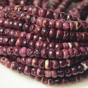 May include: Close-up of strands of dark purple beads. The beads are cylindrical with a slightly irregular shape and a glossy finish. Some beads have lighter purple and white accents. The beads are strung together, creating a layered effect.