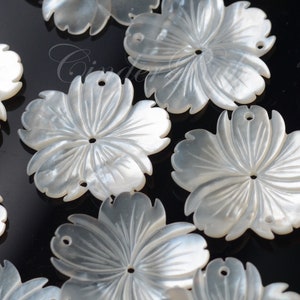 May include: A collection of white mother of pearl flower-shaped embellishments with carved details. Each flower has a hole in the center and two holes on the sides.