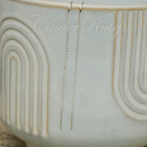 May include: A pair of silver chain earrings with a simple design. The earrings are hanging from a white ceramic bowl.