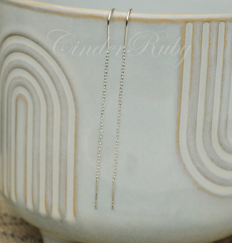 May include: A pair of silver chain earrings with a simple design. The earrings are hanging from a white ceramic bowl.
