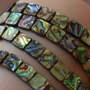 May include: A strand of iridescent green, brown, and blue abalone shell beads. The beads are square shaped and have a smooth, polished finish.