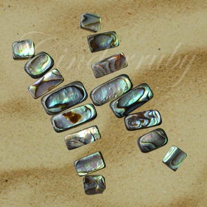 May include: A collection of iridescent abalone shell rectangles, varying in size, arranged on a light brown surface. The shells display a range of colors, including blues, greens, and browns, with a shimmering, pearlescent effect. The arrangement suggests a craft or jewelry supply.