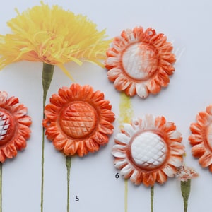 May include: Close-up of six orange and white carved sunflower pendants. The pendants have a detailed design with a textured center and petal-like edges. The background includes a yellow flower and stems.