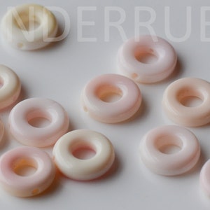 May include: A collection of 12 pink and white shell donut beads with a hole in the center. The beads are arranged in a random pattern on a white surface.