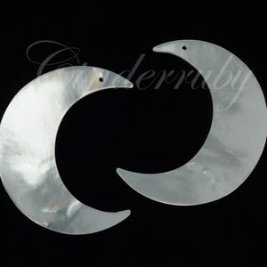 May include: Two iridescent, crescent-shaped pendants with a pearlescent sheen. Each has a small hole at the top for stringing. The pendants are set against a black background.