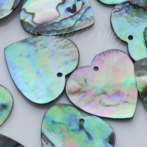 May include: A group of iridescent abalone shell heart-shaped pendants with a hole at the top for stringing. The shells have a variety of colors, including green, blue, and purple.