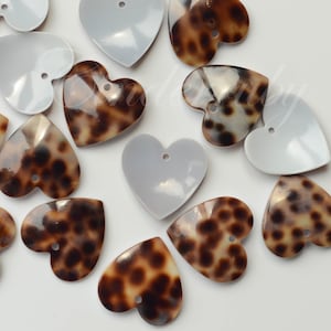 May include: A collection of heart-shaped buttons featuring a mix of solid white and brown tortoiseshell patterns. Each button has a smooth, glossy finish and a single small hole at the top for attachment, arranged in a scattered layout.
