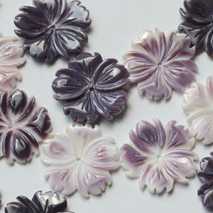 May include: Close-up of several carved flower-shaped beads in shades of purple and white. The beads have detailed petal designs and a glossy finish. These decorative beads are ideal for jewelry making and craft projects.