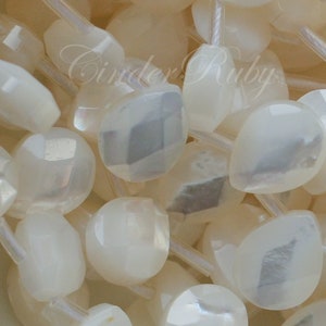 May include: A close-up of a strand of white iridescent gemstone beads. The beads are faceted and have a pear shape. The beads are strung on a white thread.