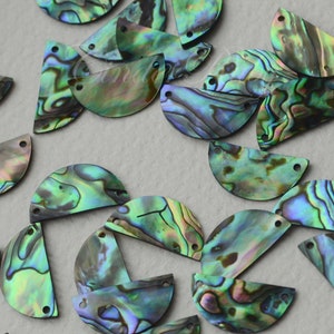 May include: A collection of iridescent green, blue, and purple abalone shell half-circles with two holes each. The shells are arranged in a random pattern on a light gray surface.