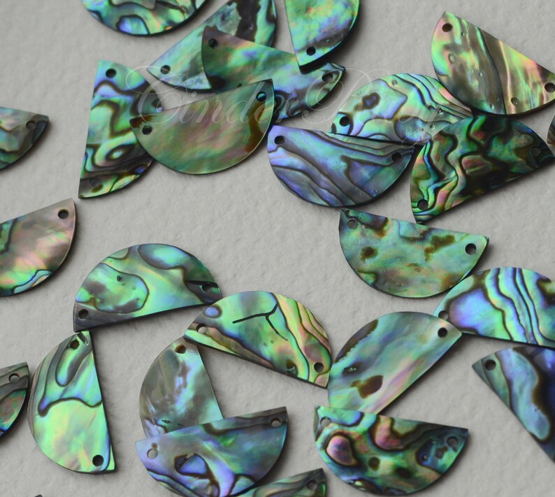 May include: A collection of iridescent green, blue, and purple abalone shell half-circles with two holes each. The shells are arranged in a random pattern on a light gray surface.