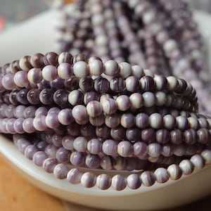 May include: A collection of round, smooth beads featuring a variegated pattern of white, light purple, and deep violet tones. The beads are strung together in multiple parallel rows, displaying a natural, polished finish with subtle color variations.