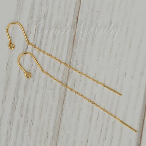 May include: A pair of gold chain earrings with a hook closure. The earrings are on a white wooden surface.