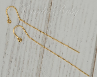 May include: A pair of gold chain earrings with a hook closure. The earrings are on a white wooden surface.