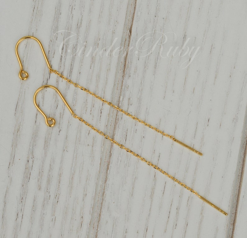 May include: A pair of gold chain earrings with a hook closure. The earrings are on a white wooden surface.