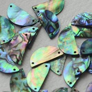 May include: A group of iridescent green, blue, and black abalone shell half-circles with two holes in each. The shells are arranged in a random pattern on a white surface.