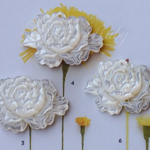 May include: Three white mother-of-pearl flower embellishments with a raised design. The flowers are arranged in a row on a white background with yellow flowers in the background.