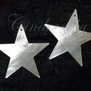 以下が含まれることがあります： 吊り下げ用の穴が上部にある、2つの真珠光沢のある白い星型のペンダント。星はきらめく虹色の品質を持ち、暗く質感のある背景に配置されています。「Cinderella」という単語がイタリック体で書かれています。