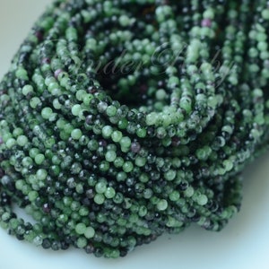 May include: A strand of green and black gemstone beads. The beads are small and round, and they are strung together on a clear thread.