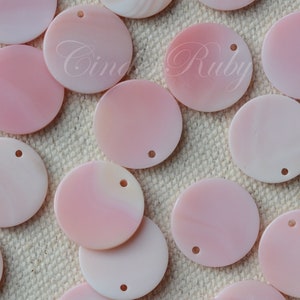 May include: A collection of round, pink, and white shell discs with a hole in the center. The discs are arranged in a staggered pattern on a beige fabric background.