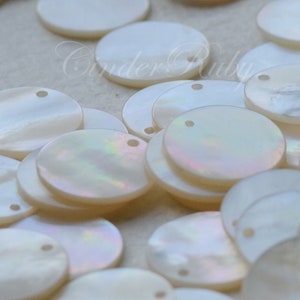 May include: A collection of white mother of pearl round discs with a hole in the center. The discs are various sizes and are scattered on a white surface.