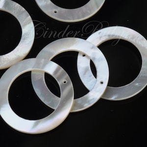 May include: A group of six white mother of pearl rings with a hole in the center. The rings are arranged in a random pattern on a black surface.