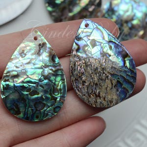 May include: Two iridescent abalone shell teardrop pendants with a hole at the top for stringing. The shells have a green, blue, and brown color pattern.