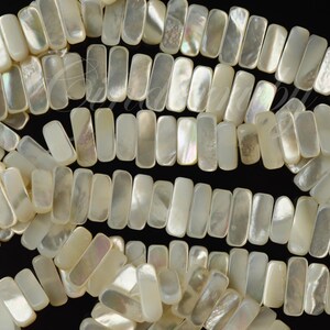 May include: Strands of rectangular, iridescent mother-of-pearl beads. The beads are a creamy white color with a subtle shimmer, arranged in rows against a black background. The beads are uniform in size and shape, ideal for jewelry making.