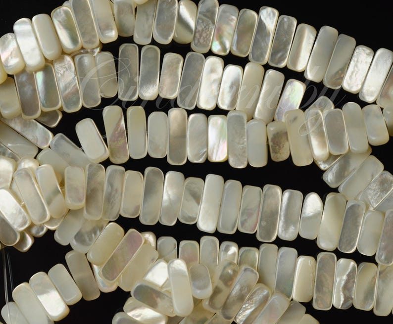 May include: Strands of rectangular, iridescent mother-of-pearl beads. The beads are a creamy white color with a subtle shimmer, arranged in rows against a black background. The beads are uniform in size and shape, ideal for jewelry making.