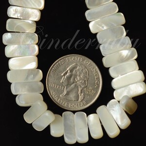 May include: Close-up of a necklace made of rectangular, iridescent, white mother-of-pearl beads. A U.S. quarter coin is placed next to the beads for size comparison. The beads are strung together, and the background is black.