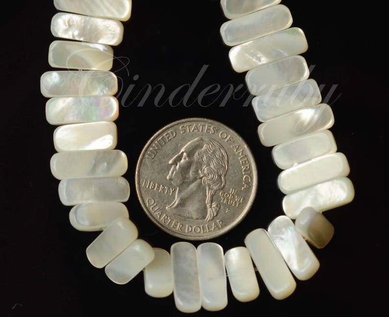 May include: Close-up of a necklace made of rectangular, iridescent, white mother-of-pearl beads. A U.S. quarter coin is placed next to the beads for size comparison. The beads are strung together, and the background is black.
