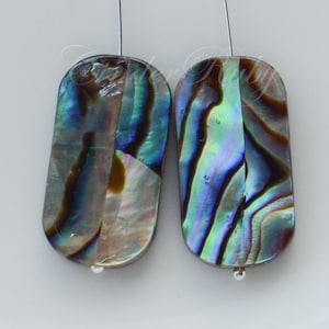May include: Two iridescent abalone shell beads with a silver wire loop at the top. The beads are oval shaped and have a blue, green, and brown color pattern.