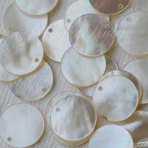 Peut inclure: Une collection de disques ronds en nacre blanche et irisée avec un trou au centre. Les disques sont disposés de manière aléatoire sur une surface blanche.