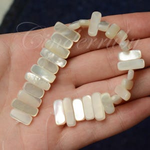 May include: A strand of rectangular, iridescent mother-of-pearl beads. The beads are strung together, forming a flexible chain. The beads have a creamy white color with subtle variations in tone. The overall theme is jewelry and crafting.