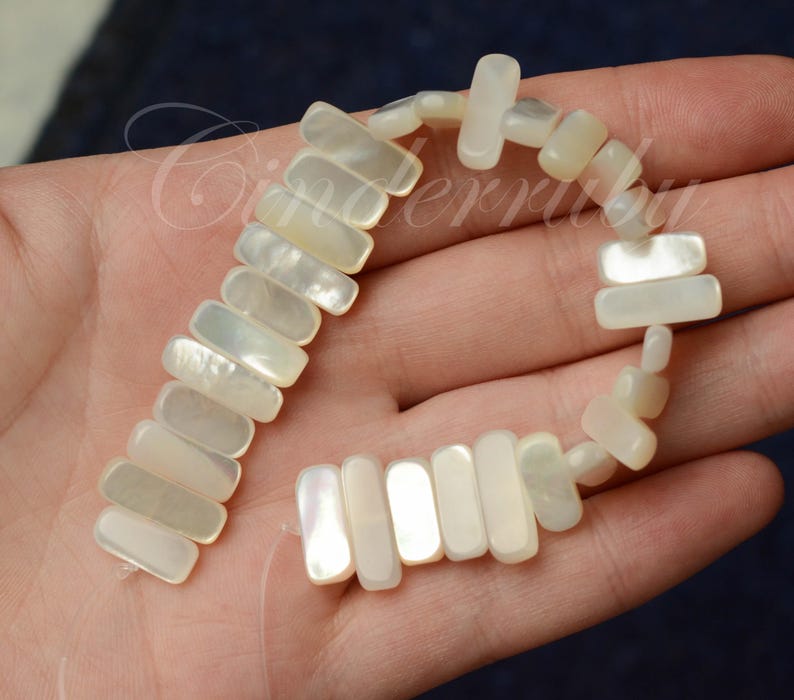 May include: A strand of rectangular, iridescent mother-of-pearl beads. The beads are strung together, forming a flexible chain. The beads have a creamy white color with subtle variations in tone. The overall theme is jewelry and crafting.