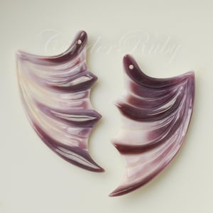 May include: Two wing-shaped pendants in shades of purple and white. Each pendant has a hole at the top for stringing. The wings have a layered design with curved edges, creating a textured appearance. The background is a plain, off-white surface.