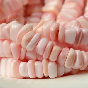 May include: A collection of smooth, polished, irregular-shaped beads in varying shades of soft pastel pink and creamy white. The beads are arranged in multiple rows, showcasing a glossy, reflective surface and a rounded, organic texture throughout.