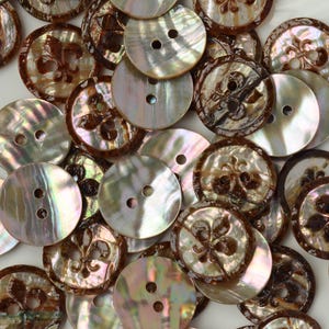 May include: A collection of round, iridescent mother-of-pearl buttons in shades of brown and cream. Some buttons feature a two-hole design, while others are engraved with a brown fleur-de-lis pattern. The surface has a glossy, pearlescent finish.
