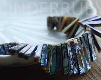 May include: A strand of iridescent abalone shell beads, each bead is a long, thin rectangle with a natural, organic shape. The beads are arranged in a curved line, creating a flowing, elegant look.