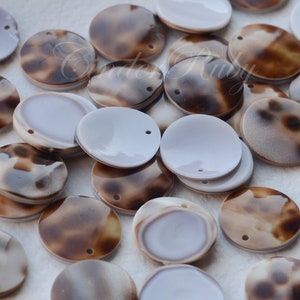 May include: A collection of round, brown and white shell-like discs with a hole in the center. The discs are arranged in a random pattern on a white surface.